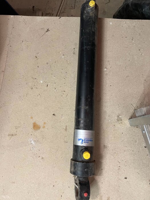 Hydraulcylinder på ett bord, tidigare ägares överblivna objekt från husköp. Svart med gula detaljer och etikett, potentiellt för försäljning.