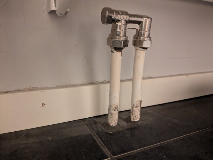 Två vita rör med metallkopplingar sticker upp ur golvet nära en vit vägg och sockel.