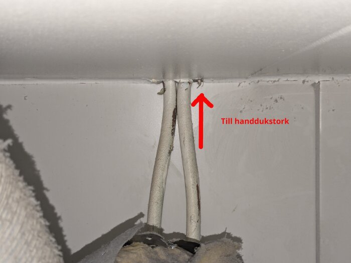 Två vita rör går in i taket med en röd pil och texten "Till handdukstork" bredvid. De är delvis isolerade och omgivna av isoleringsmaterial.