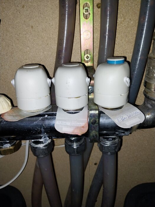 Tre vita termostatventiler monterade på rör, märkta med etiketter för olika zoner, mot en träpanel.