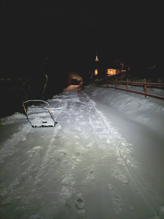 Upplyst snötäckt väg med gångstig, snöruta och spår nära ett upplyst hus på kvällen.