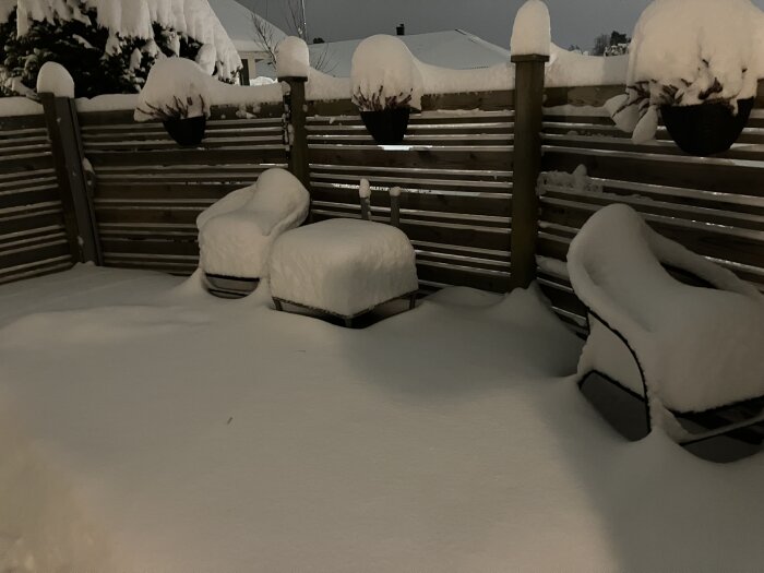 Snötäckt uteplats med stolar, bord och staket under tjockt lager av snö, kvällstid.