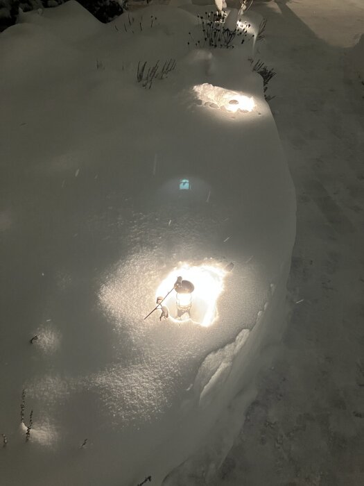 Trädgårdsbelysning under snö, lampor omgivna av snötäcke med små insjunkna områden runt ljuspunkterna, mörk omgivning.