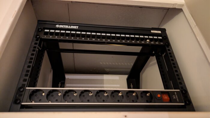 Ett litet svart rack på väggen med en patchpanel för ethernetkablar, placerat i ett ventilerat överskåp med eluttag synliga.