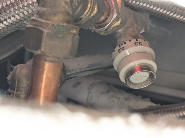 Närbild av en strypventil med ett reglage markerat vid 0.5, omgiven av kopparrör och metallslangar.