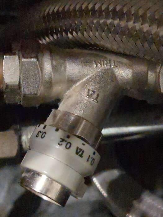 En närbild av en metallventil med präglad text "TA" och "1/2", samt ett reglage med markeringarna "0.1", "0.2", och "0.3".