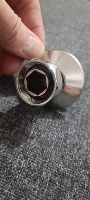 Metallisk kabinkoppling för duschblandare, med gängat inlopp och hexagonalt fäste, hålls av en hand på grå texturerad yta.