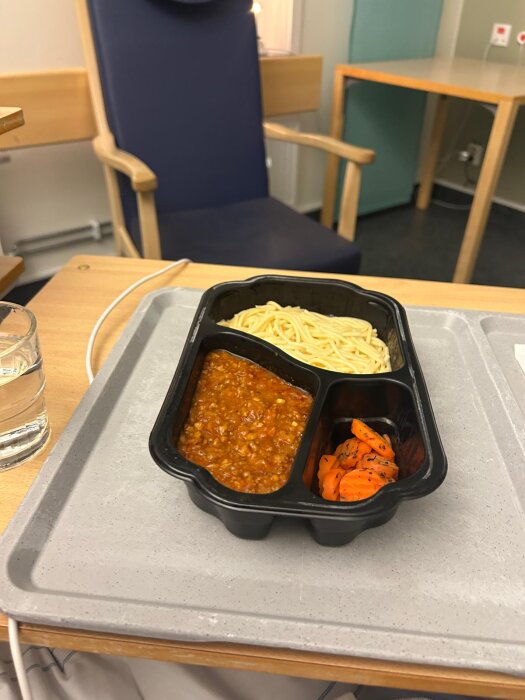 Plastlåda med pasta, köttfärssås och morotsbitar på en sjukhusbricka, placerad i en vårdmiljö.