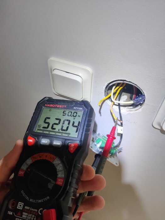 En multimeter visar 52,04V AC framför en vägguttag med kablar exponerade; koppling och mätning av mellantrådar syns.