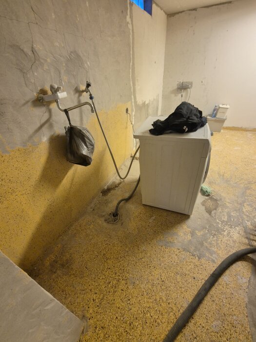Gult golv med golvbrunn och tvättmaskin, vägg med exponerad betong och rör.