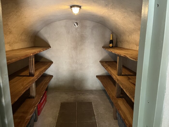 Jordkällare med trähyllor och en flaska på översta hyllan, lampa i taket.