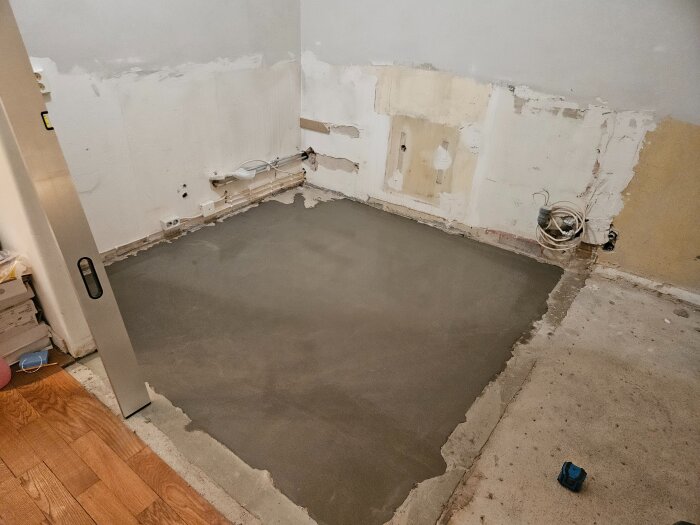 Rum under renovering med nylagt flytspackel på golvet, omgivande väggar med uppbrutet gips och träplankor till vänster.