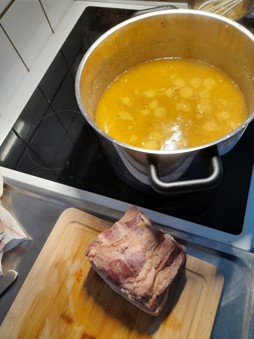 Gryta med våt ärtsoppa på spis och köttbit på skärbräda bredvid.