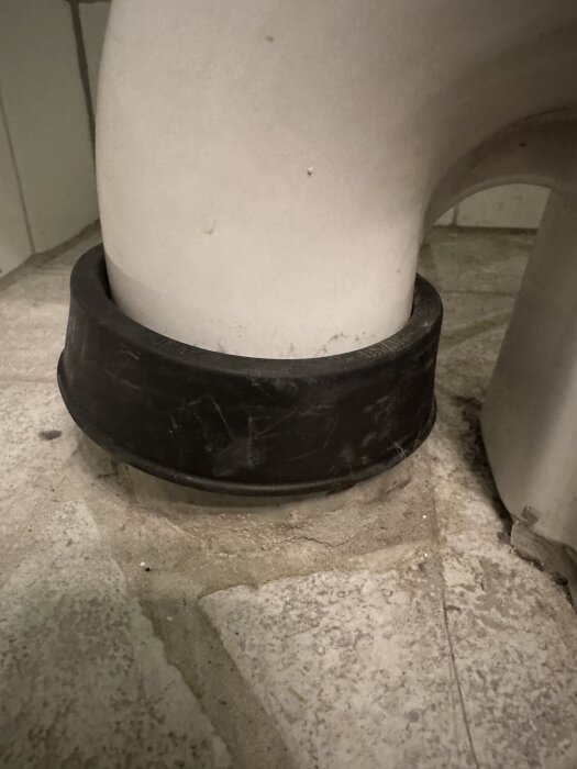 Gammalt toalettanslutningsrör med svarta wc-manschetten synligt, omgiven av kakelgolv.
