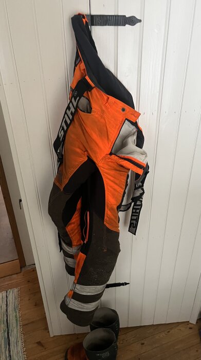 Arbetsbyxor i orange och svart, märkta Stihl, hänger på en vit trävägg. Passar användning med röjsågning i kallt väder.