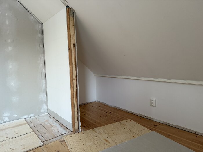 Övervåning med synlig träregel och spånskiva bakom gipsvägg, delvis blottat område visar ombyggnation. Golv av trä och plywood. Vägguttag syns.