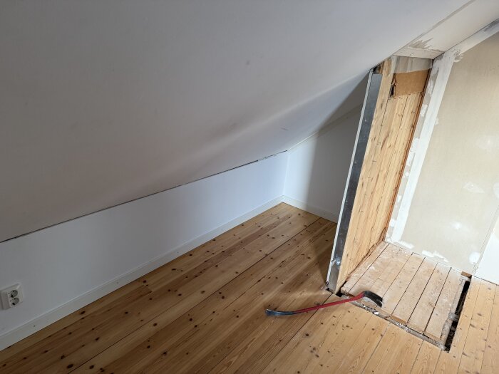 Rum med snedtak, träpanel och gipsskiva, en delvis riven vägg med träreglar syns. Ett brytjärn ligger på golvet av trägolv.