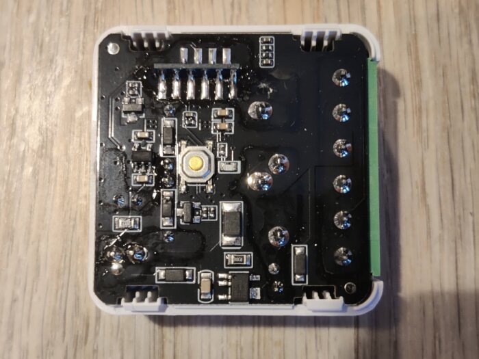 Elektronisk krets med flera komponenter på ett fyrkantigt kretskort, inuti en vit plastbehållare på ett träbord.