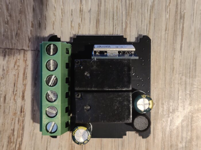 Elektronisk modul med grön terminalblock, två svarta reläer och kondensatorer monterad på ett kretskort med trämönstrad bakgrund.