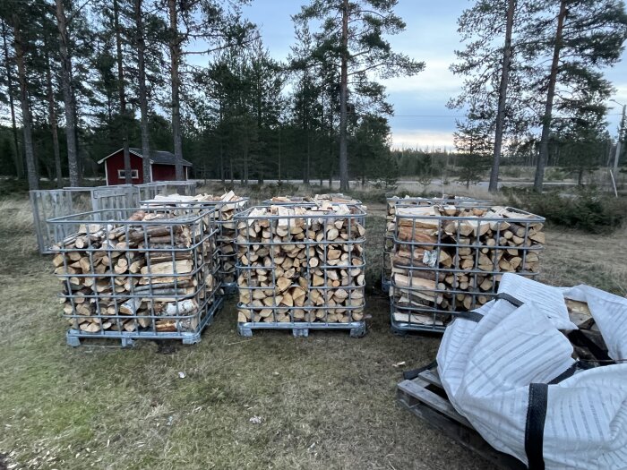 Tre staplar med ved i metallburar utomhus, omgivna av skog och en röd stuga i bakgrunden.