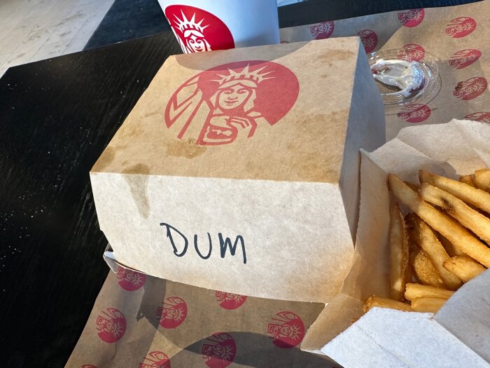 En bricka på ett matställe med en umami-burgare i kartong märkt "DUM", pommes frites och en läskmugg i bakgrunden.