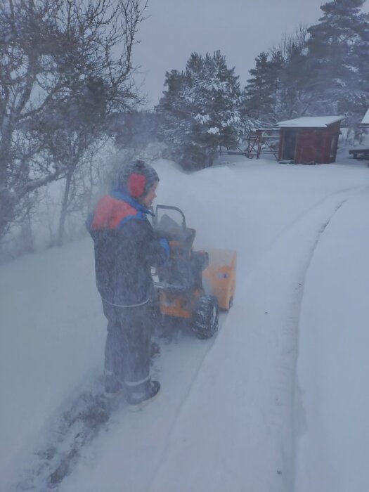 Person använder snöslunga för att röja snö en vinterdag, omgiven av snötyngda träd.