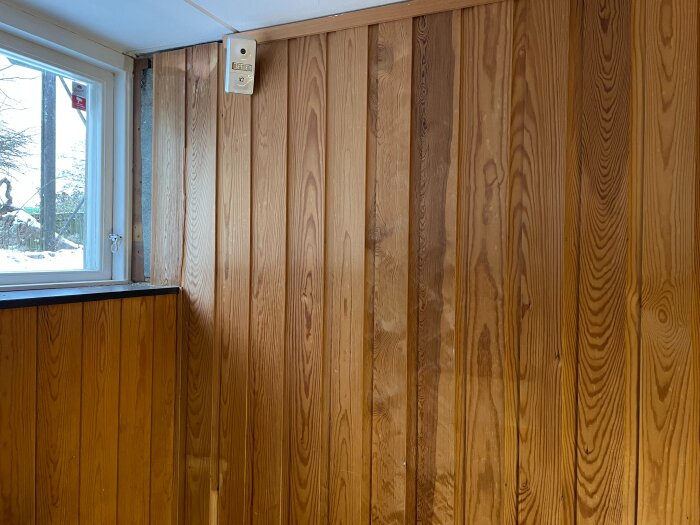 Träpanel på vägg i källare med ett litet fönster och en ventil. En sektion av panelen är borttagen för inspektion.