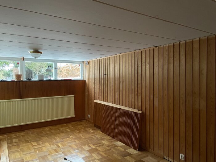 Gillestuga med träpaneler på väggarna, parkettgolv, och fönster med utsikt mot trädgård. Element placerat längs väggen.
