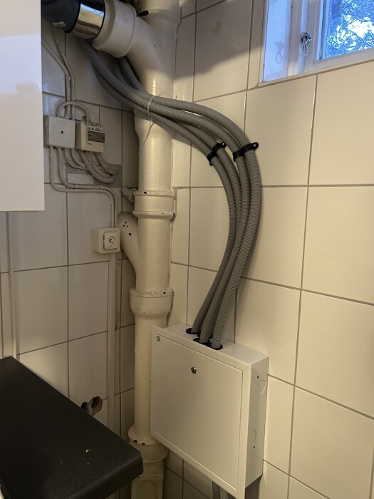 Grå rör-i-rör installation i ett kaklat tvättstugerum, kopplat till kopplingsskåp under diskbänken genom taket från ett badrum ovanför.