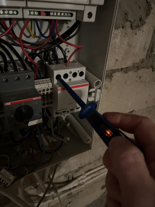 En hand håller en spänningsprovare över en elektrisk brytare i en kopplingsdosa, omgiven av olika kopplade kablar.