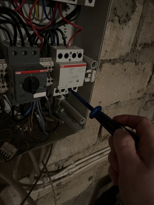 En hand använder en spänningsprovare på en elektrisk brytare i en elcentral med flera kablar anslutna.