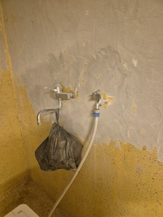 Vägg i tvättstuga med två blandare, silverfärgad kran och en svart plastpåse upphängd. Sliten gul och grå vägg med sprickor.