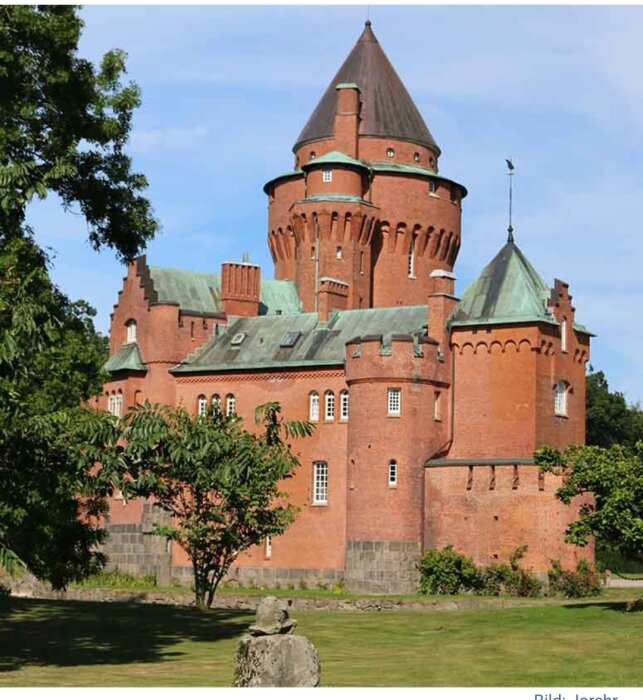 Rött tegelstensslott med torn och grönt tak, omgiven av träd och gräsmatta, under en klarblå himmel.
