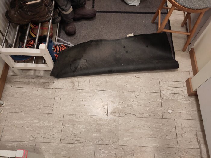 En plastmatta ligger delvis utrullad över ett stengolv intill en skoställning med skor.