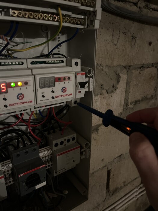 Elektrisk fördelningslåda med synliga kablar där en person använder en spänningsprovare mot en kopplingspunkt i en tegelbyggnad.