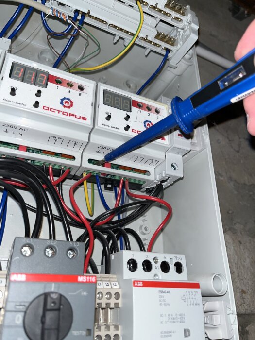 Elektrisk kopplingsbox med LED-displayer och statuslampor. Verktyg pekar på en kontakt, omgiven av kablar och anslutningar.