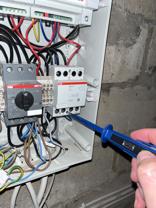 Person använder skruvmejsel på elektrisk kopplingslåda med flera färgade kablar, mot stenmur i bakgrunden.