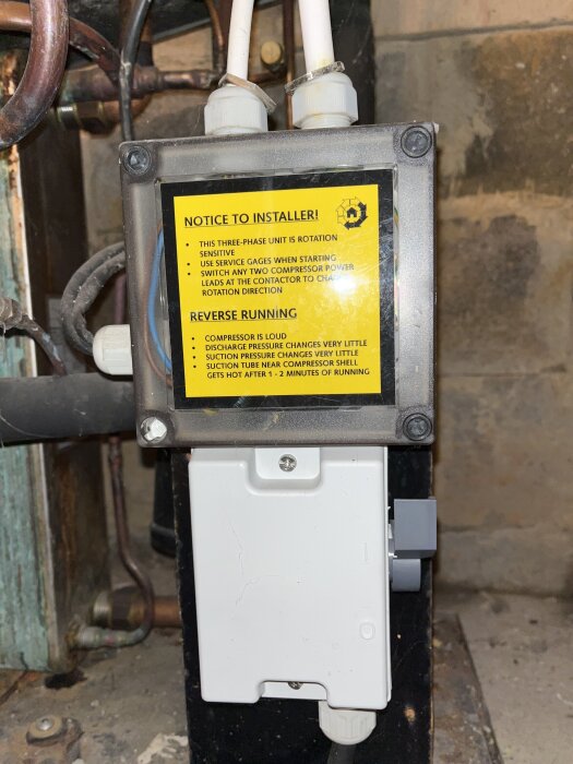 Elektrisk kontrollbox med gul varningsetikett och rörkopplingar ovanför, omgiven av kablar och metallrör i en teknisk installation.
