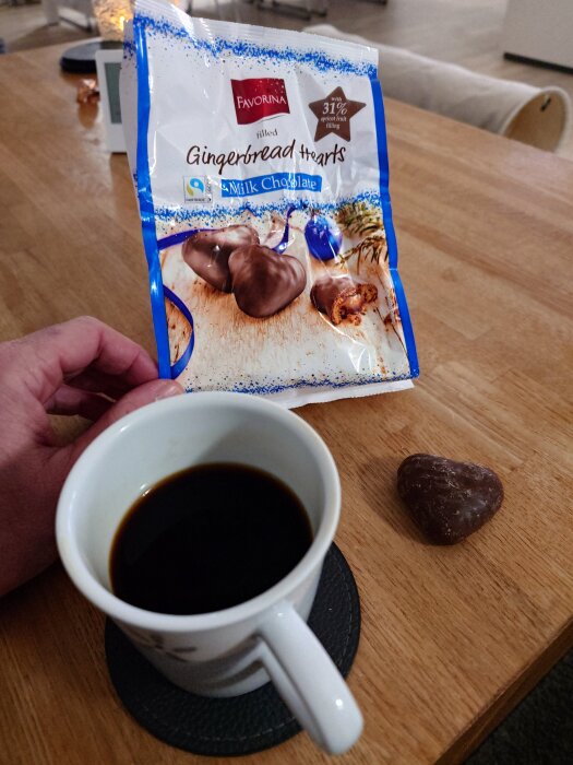 En hand håller en påse med chokladhjärtan bredvid en kopp svart kaffe och ett chokladhjärta på ett träbord.