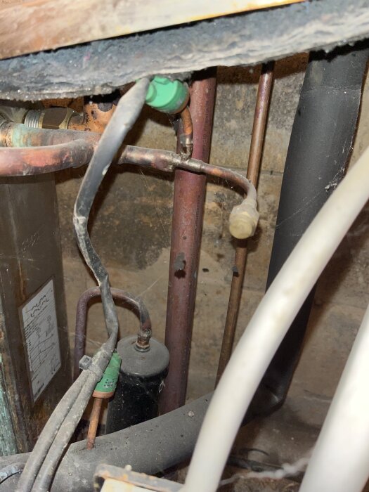 Rostiga kopparrör och kablar i en trång utrymme med green komponenter, en svart cylinder och en etikett på en metallbox i bakgrunden.