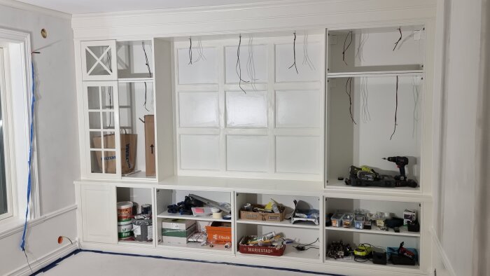 Platsbyggd vit hylla och TV-bank under renovering, med verktyg och material på hyllorna. Oexponerade kablar hänger från väggen.