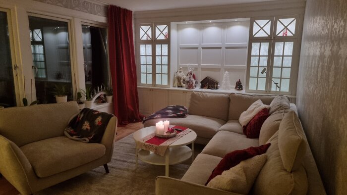 Vardagsrum med platsbyggd hylla, vitrinskåp och vägghängd TV-bank. Soffa, röd gardin, och tända ljus på bordet. Fönster med juldekorationer.