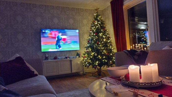 Vardagsrum med julgran och tända ljus, TV på väggen visar sport, soffbord med ljus och godis, rummet nyrenoverat och inrett.