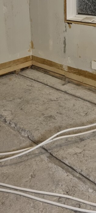 Rivet golv i ett rum, exponerar den underliggande cementytan och kablar, med märken efter borttagen våtrumsmatta på väggarna.