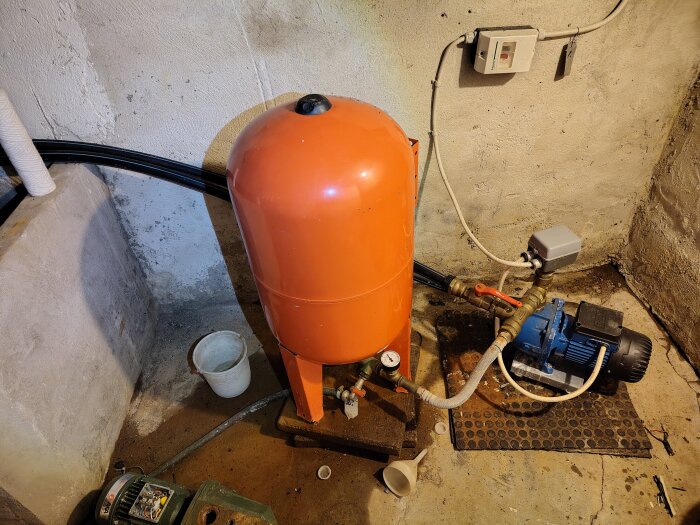 Orange hydrofor med rör och blå vattenpump i betongrum. Eluttag på väggen och plastmugg står bredvid.