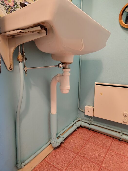 Slitet handfat i fritidshus med röranslutning och blått avloppsrör mot turkos vägg, samt orange kakelgolv och element i bakgrunden.