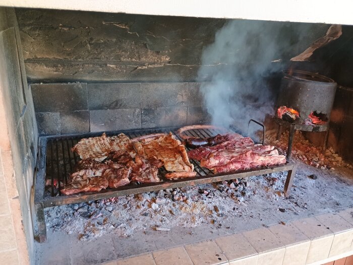 Grillad kött och korv på utegrill med rök, vedeldning på sidan, aska under gallret.