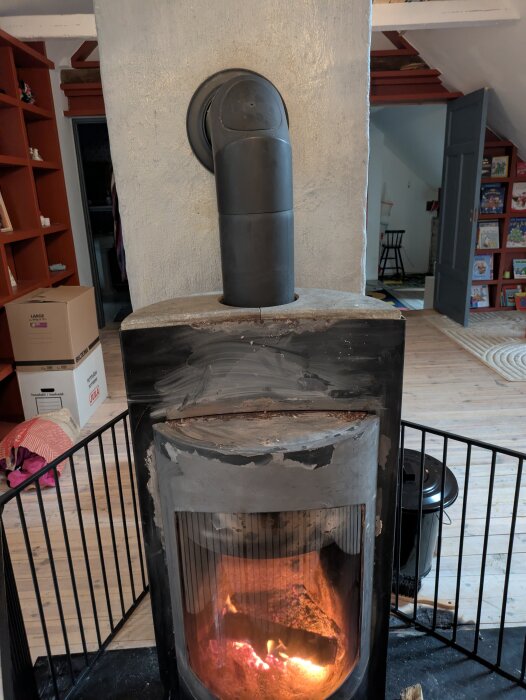 Vedspis med glaslucka och synlig brasa, omgiven av säkerhetsgaller. Rött tegel och bruna möbler i bakgrunden.
