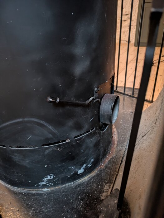 Kamin med en spjällmekanism på sidan, del av asklåda synlig och sotmärken runt kanten. Inomhusgolv av trä i bakgrunden.