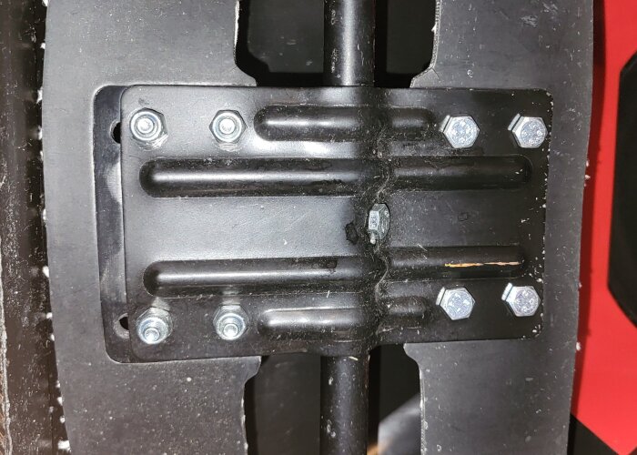 Svart metallplatta monterad med åtta silverfärgade bultar på en svart yta.